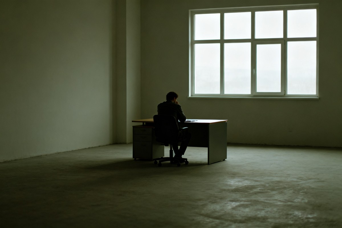 Personne seule assise pensivement à un bureau près d'une fenêtre dans un open space sombre