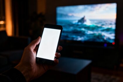 Un smartphone allumé dans un salon sombre, un grand écran de télévision éclairé en arrière-plan.