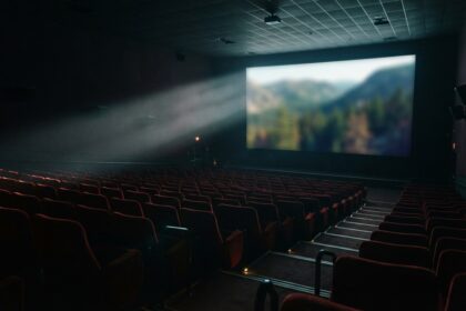 Salle de cinéma vide aux fauteuils rouges, un faisceau de projection éclairant un écran où passe un paysage de montagne