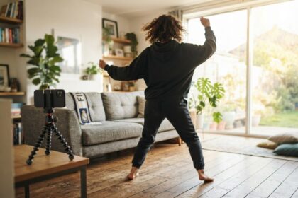 Une personne de dos danse dans un salon lumineux, filmée par un smartphone sur trépied, une écharpe de foot sur le canapé