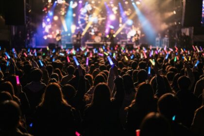 Foule de concert vue de dos, mains levées tenant des bâtons lumineux colorés devant une scène éclairée.
