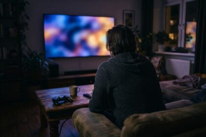 Une personne vue de dos sur un canapé, face à un grand écran allumé dans un salon plongé dans l'obscurité.