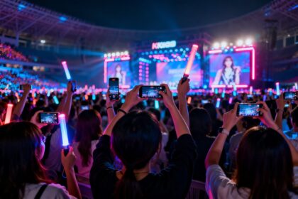 Foule de spectateurs de dos filmant un concert de K-pop au smartphone, bâtons lumineux levés sous des néons.