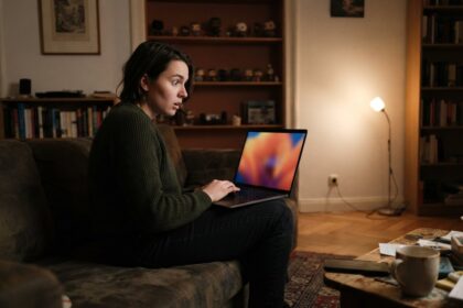 Une femme surprise regarde une vidéo sur un ordinateur portable dans un salon éclairé par une lampe, le soir.